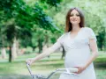 Uśmiechnięta kobieta w ciąży w białej sukience trzyma kierownicę roweru w parku.