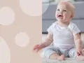 Uśmiechnięte dziecko w białej koszulce i pasiastych spodenkach siedzi na dywanie. Wskazówki, jak nauczyć dziecko siadać.