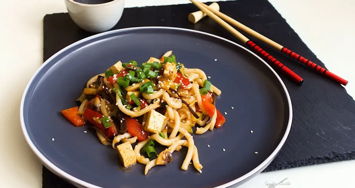 Pyszne udon z tofu, warzywami i sezamem, podane na ciemnym talerzu z pałeczkami.