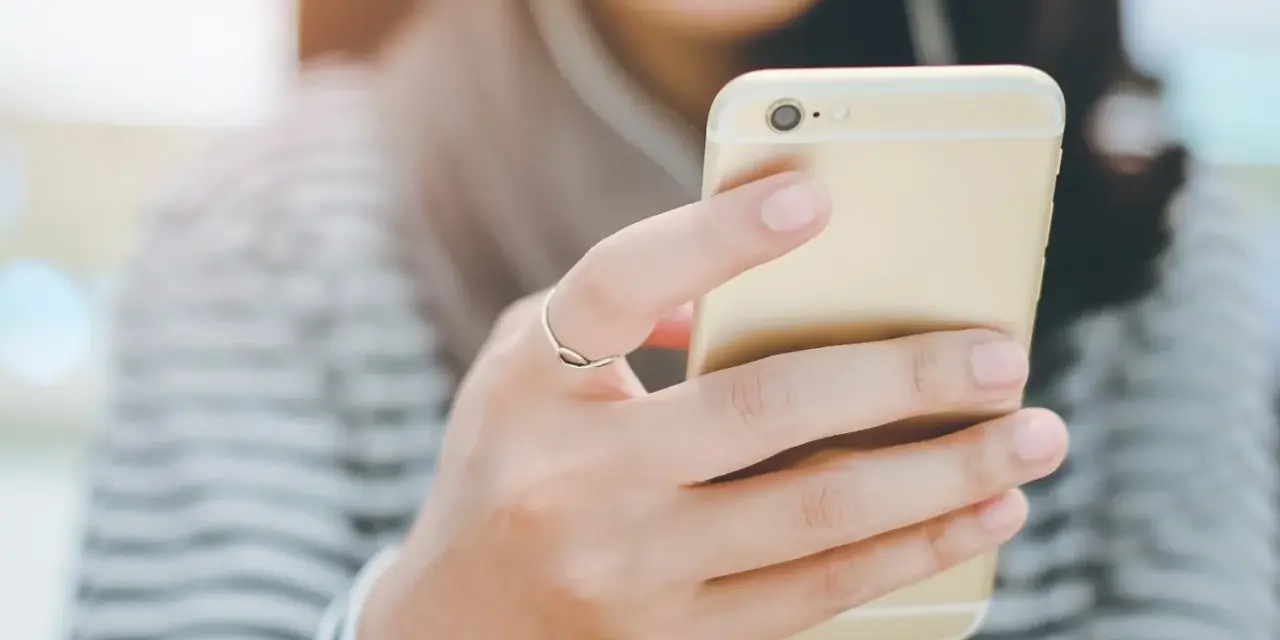 Uśmiechnięta osoba trzymająca złoty smartfon z podłączonymi słuchawkami dousznymi. Czy słuchawki douszne są szkodliwe?