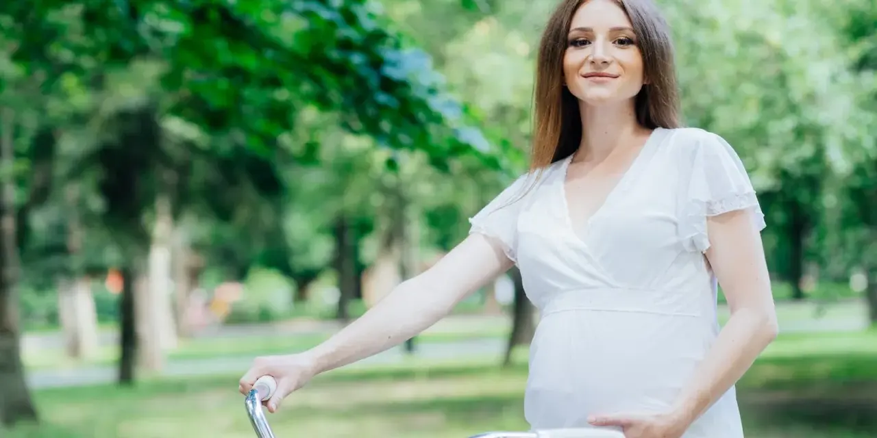 Uśmiechnięta kobieta w ciąży w białej sukience trzyma kierownicę roweru w parku.