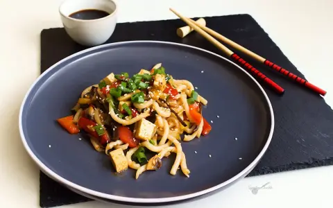 Pyszne udon z tofu, warzywami i sezamem, podane na ciemnym talerzu z pałeczkami.