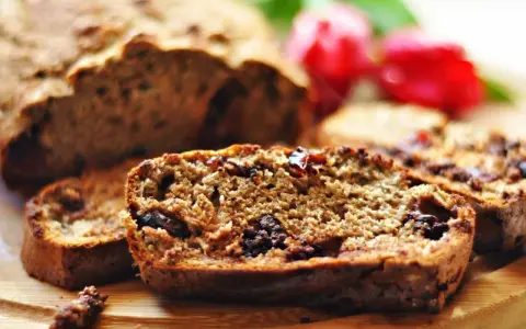 Pokrojony niskokaloryczny chlebek bananowy z kawałkami czekolady i rodzynkami, na drewnianej desce, w tle czerwone kwiaty.