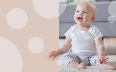 Uśmiechnięte dziecko w białej koszulce i pasiastych spodenkach siedzi na dywanie. Wskazówki, jak nauczyć dziecko siadać.
