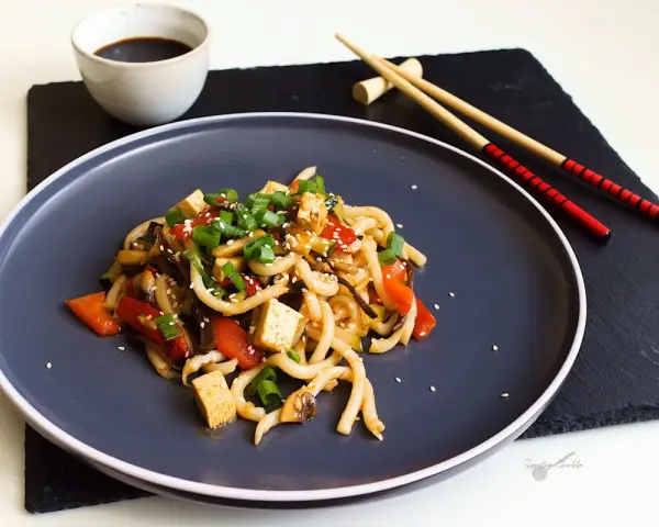 Pyszne udon z tofu, warzywami i sezamem, podane na ciemnym talerzu z pałeczkami.