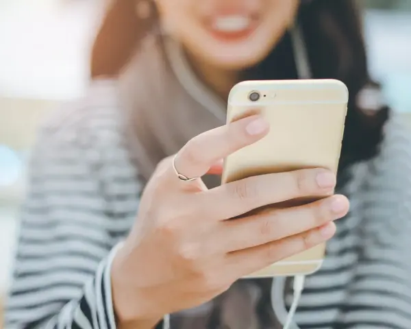 Uśmiechnięta osoba trzymająca złoty smartfon z podłączonymi słuchawkami dousznymi. Czy słuchawki douszne są szkodliwe?
