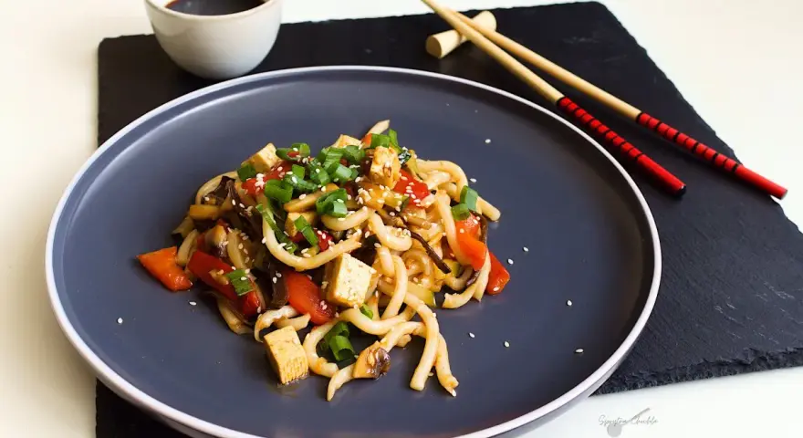 Pyszne udon z tofu, warzywami i sezamem, podane na ciemnym talerzu z pałeczkami.