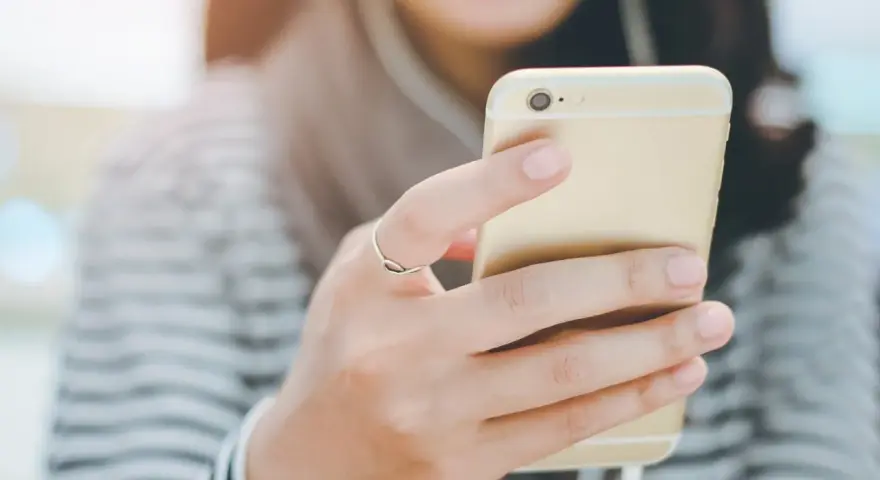 Uśmiechnięta osoba trzymająca złoty smartfon z podłączonymi słuchawkami dousznymi. Czy słuchawki douszne są szkodliwe?
