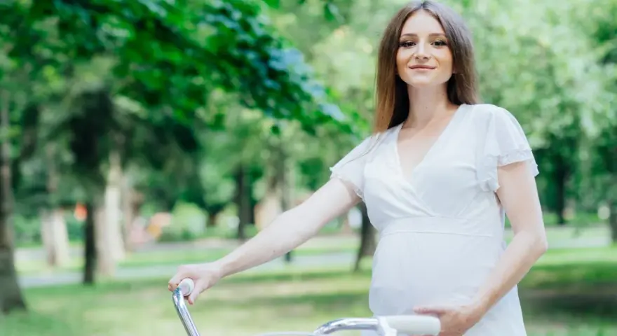 Uśmiechnięta kobieta w ciąży w białej sukience trzyma kierownicę roweru w parku.