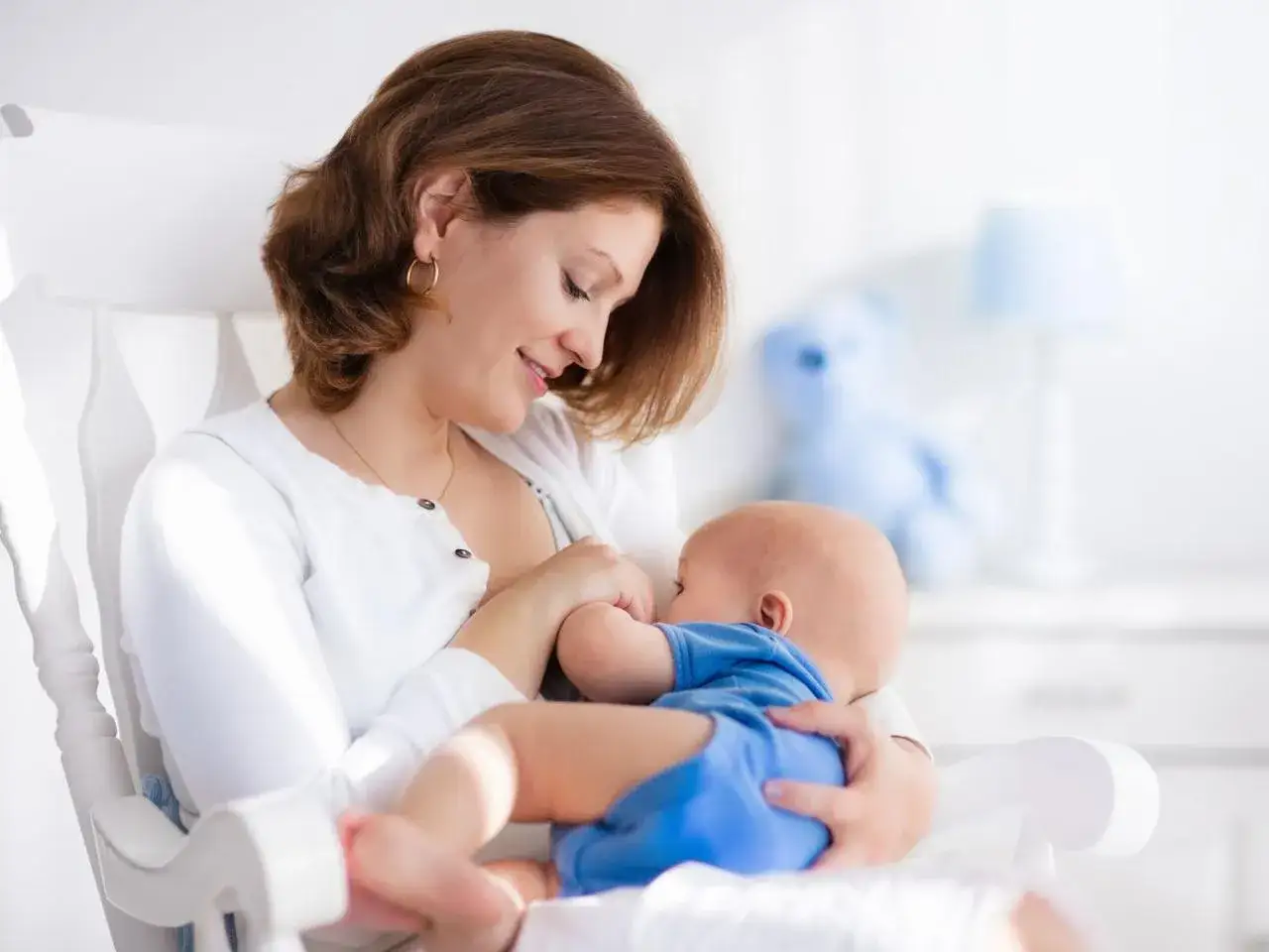 Cierpliwa mama karmi piersią, choć noworodek nie chce jeść. Uśmiecha się do maluszka, kołysząc go w fotelu.
