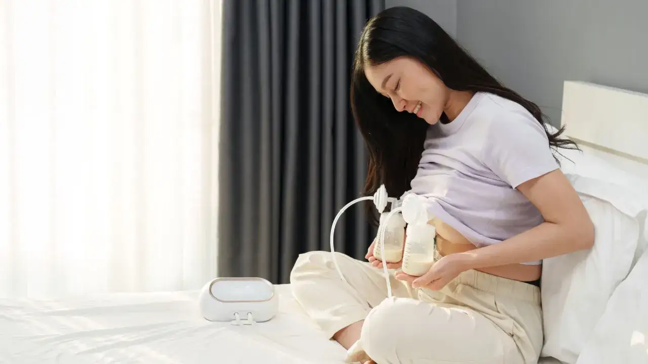 Uśmiechnięta mama pokazuje, jak odciągać mleko laktatorem. Na ł&oacute;żku siedzi kobieta z podłączonym laktatorem, zbierając mleko do butelek.