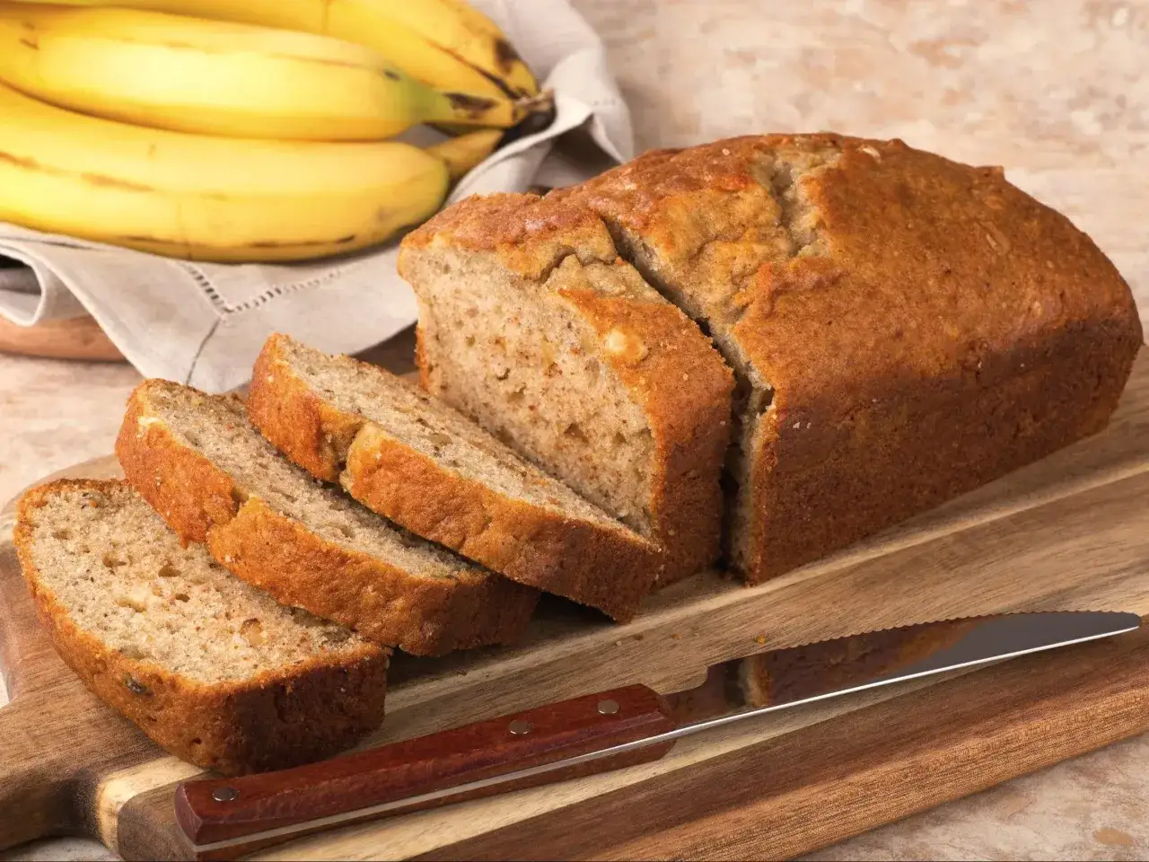 Niskokaloryczny chlebek bananowy, pokrojony w plastry, leży na desce obok pęczka bananów.